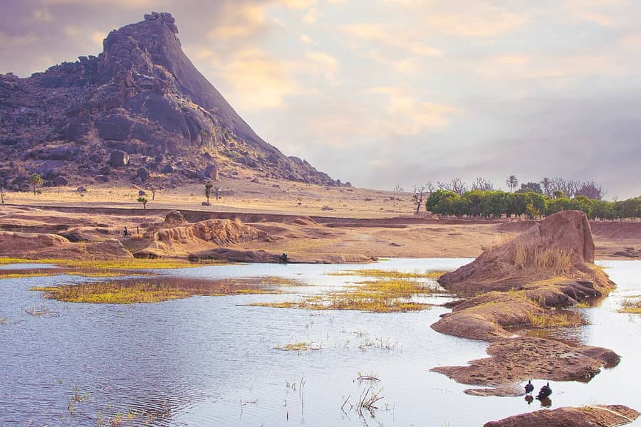 Une colline rocheuse au-dessus de la savane sèche et d'un point d'eau, dans l'État de Bauchi, au Nigeria : le pays du proverbe haoussa.