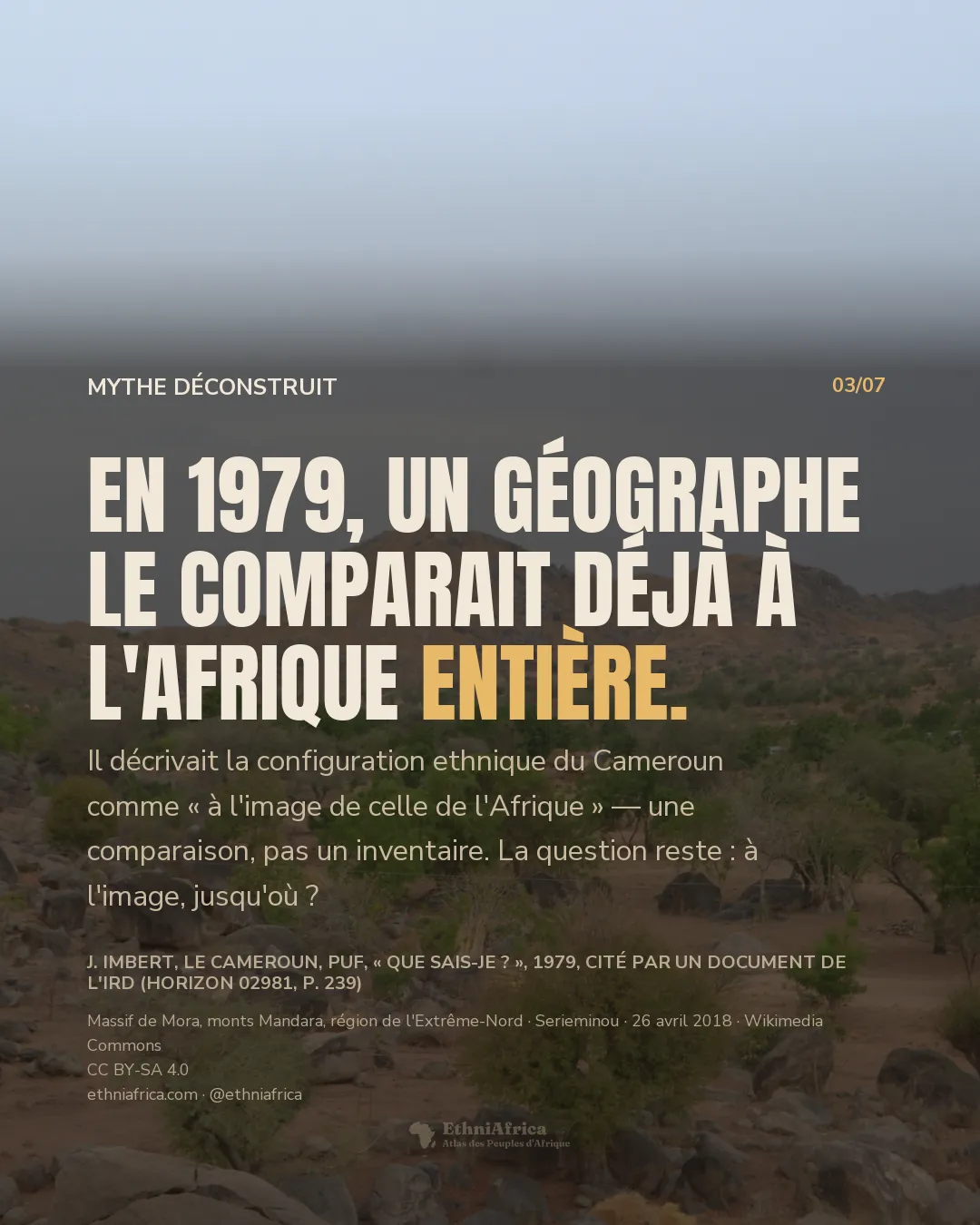 une vallée montagneuse en saison sèche dans le nord du Cameroun, avec des collines et des blocs de roche volcanique arrondis au premier plan, une végétation clairsemée et de petites cases rondes à toit de chaume regroupées parmi les arbustes, sous un ciel pâle et brumeux